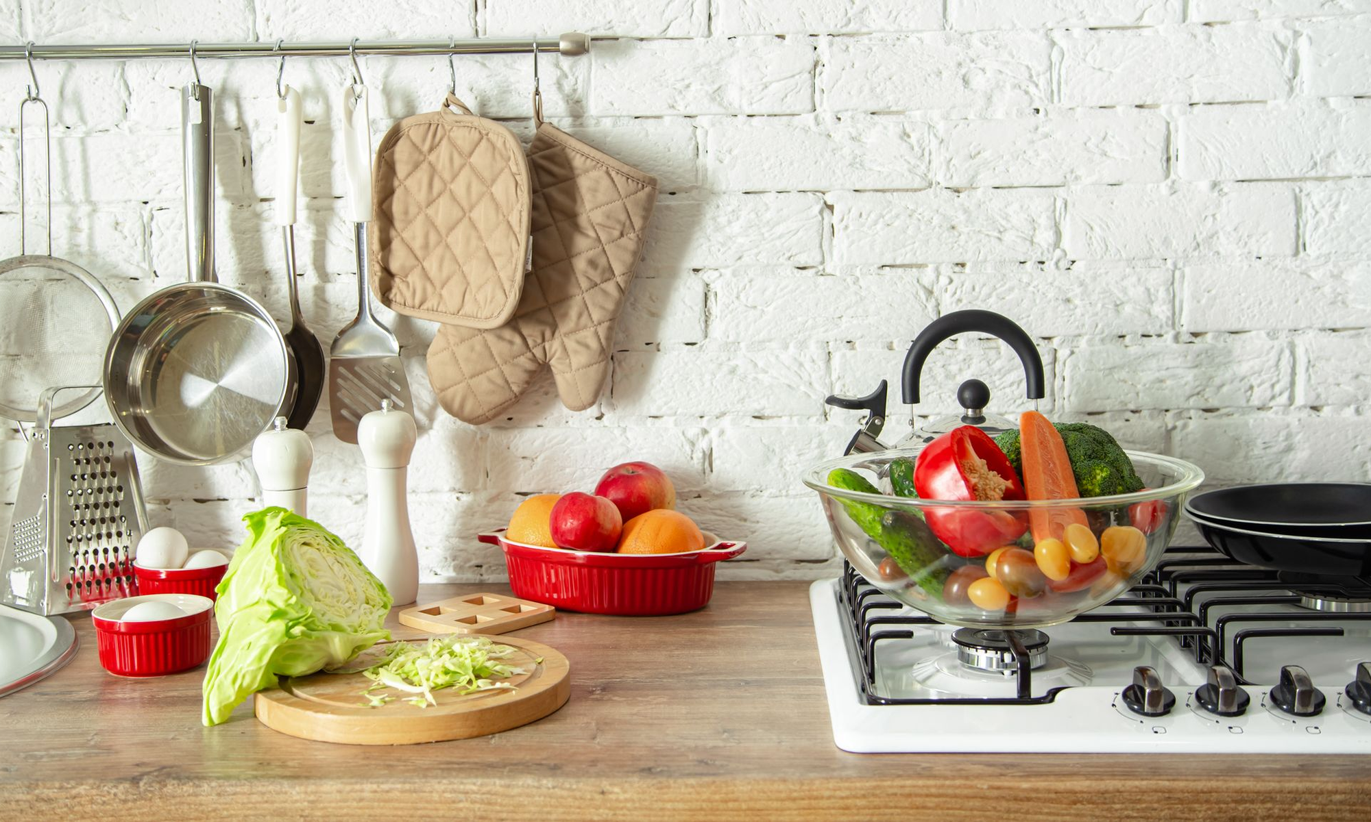 Kitchen counter with vegetables, fruits, and kitchen utensils on a white tiled wall background